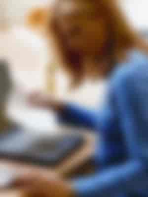Frau am Schreibtisch im blauen Pullover vor Laptop mit Zettel in der rechten Hand und linke Hand auf dem Taschenrechner