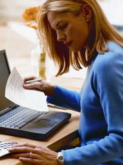 Frau am Schreibtisch im blauen Pullover vor Laptop mit Zettel in der rechten Hand und linke Hand auf dem Taschenrechner Frau am Schreibtisch im blauen Pullover vor Laptop mit Zettel in der rechten Hand und linke Hand auf dem Taschenrechner